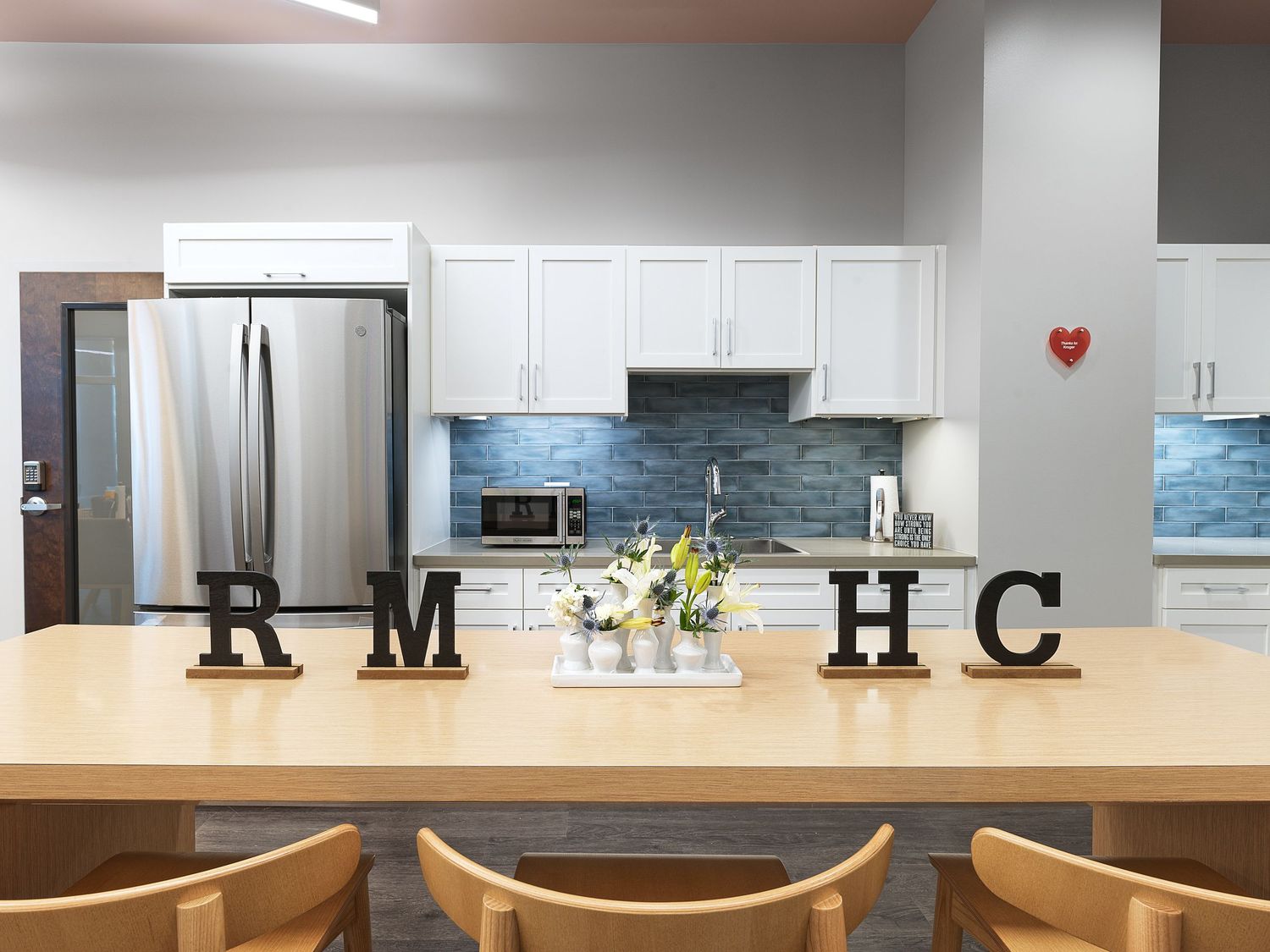 Bright kitchen dining area with white cabinets, stainless fridge, wood table, and RMHC letter decor.