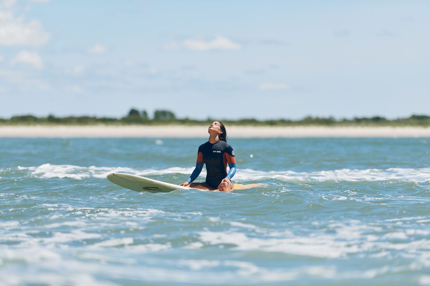 Woman in wetsuit sitting on surfboard in calm ocean waves on clear day.