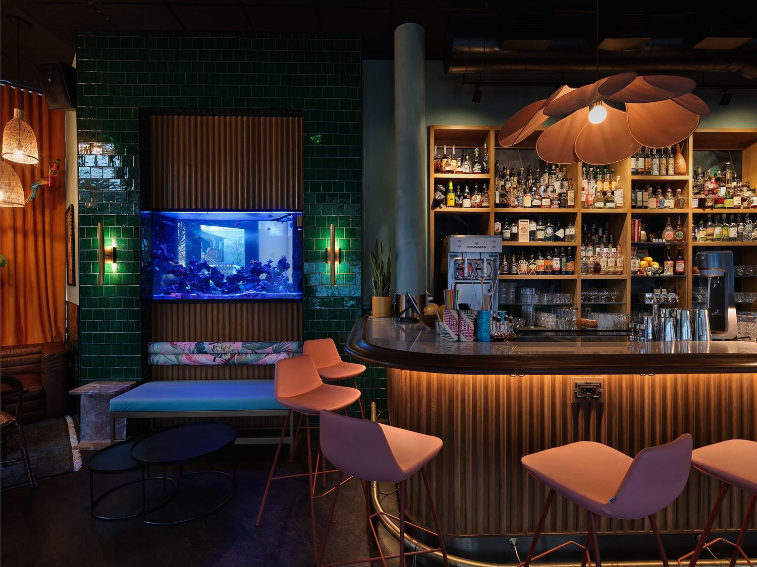 Stylish cocktail bar interior with green tile walls, fish tank, and pink bar stools.