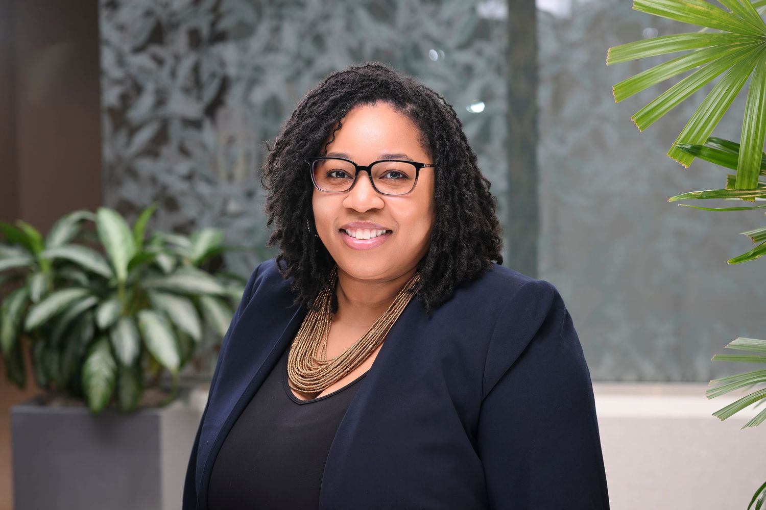 Professional woman with glasses and curly hair in navy blazer smiles in modern office with green plants.