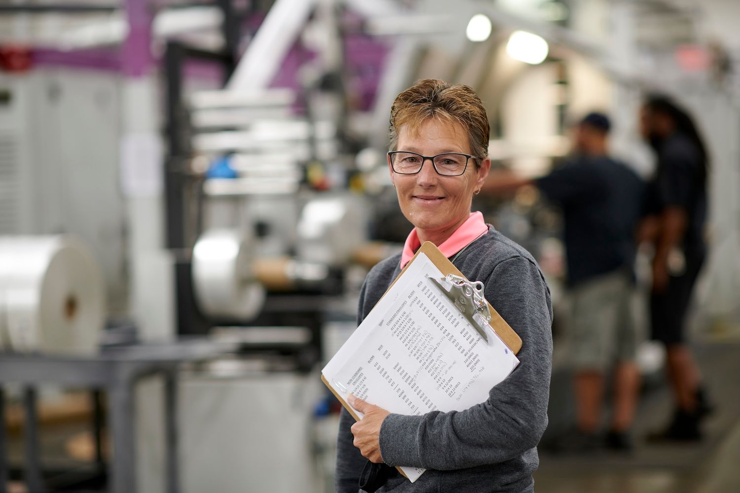 Woman holding clipboard smiling in industrial printing factory workplace.