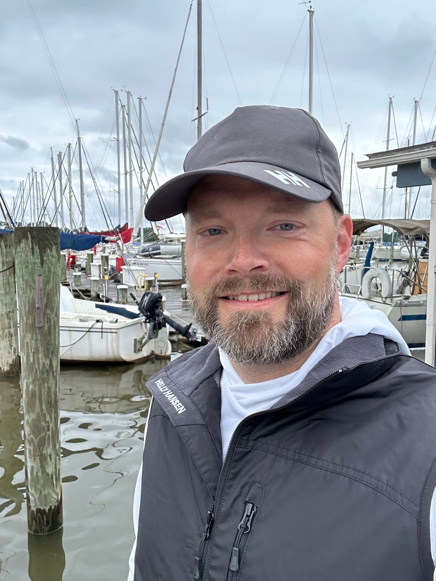 Man in gray jacket and cap smiling at a marina with sailboats in background.