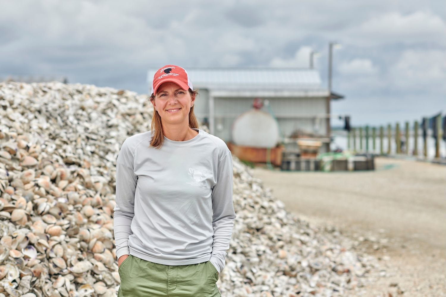 Smiling woman in red cap standing outdoors near pile of oyster shells.
