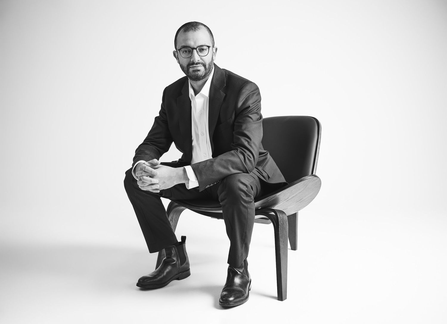 Monochrome professional photo of suited bearded man sitting on chair in minimalist setting.