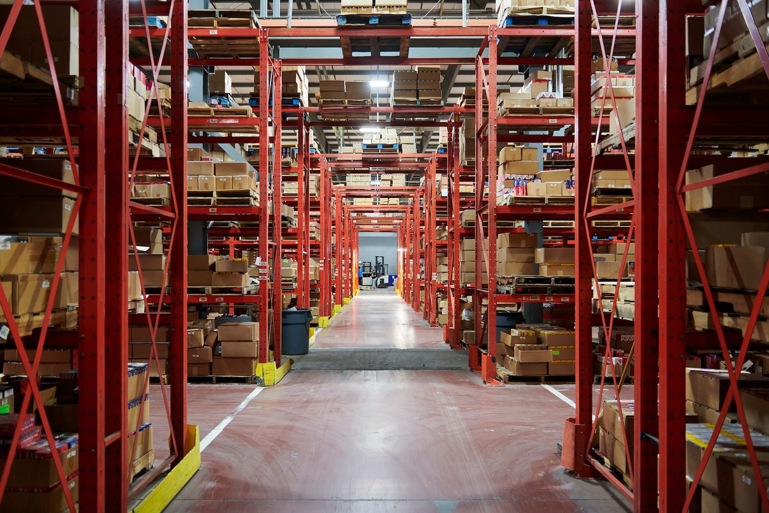 Vast warehouse interior with tall red metal shelving racks filled with boxed inventory and goods.