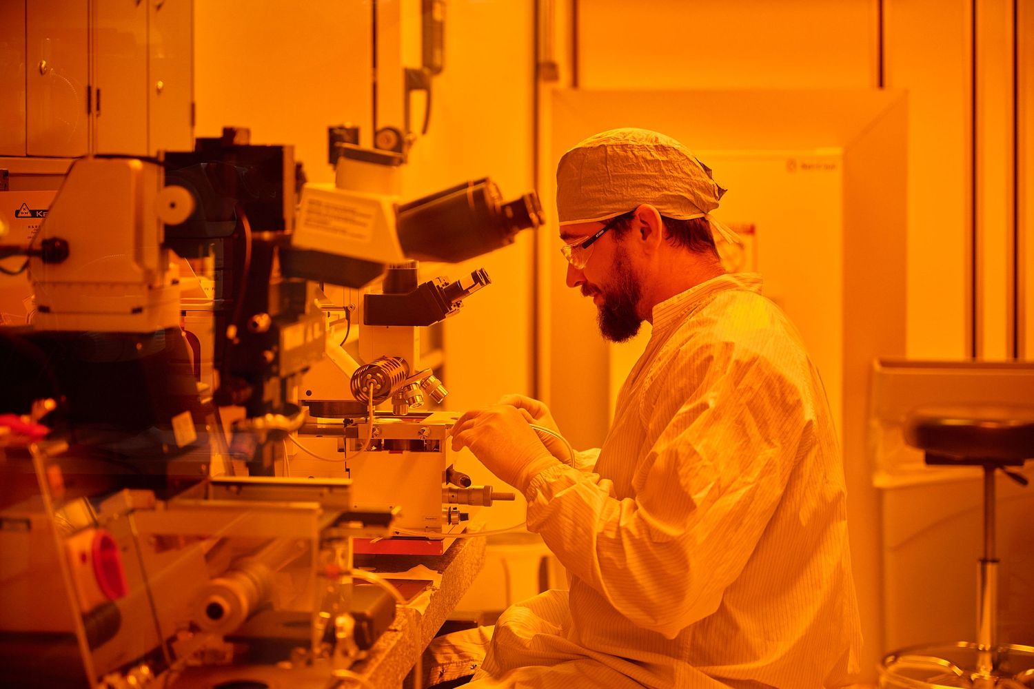 Scientist in cleanroom protective gear working under orange safelight in semiconductor research laboratory.