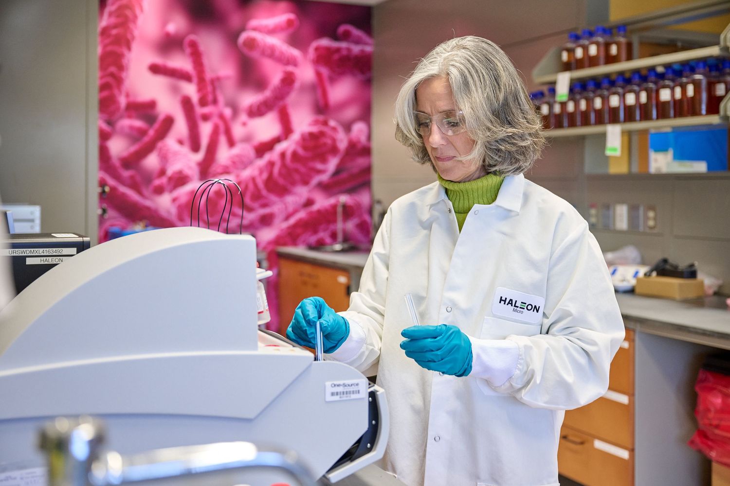 Female scientist in white lab coat using laboratory equipment with bacterial imagery in background.