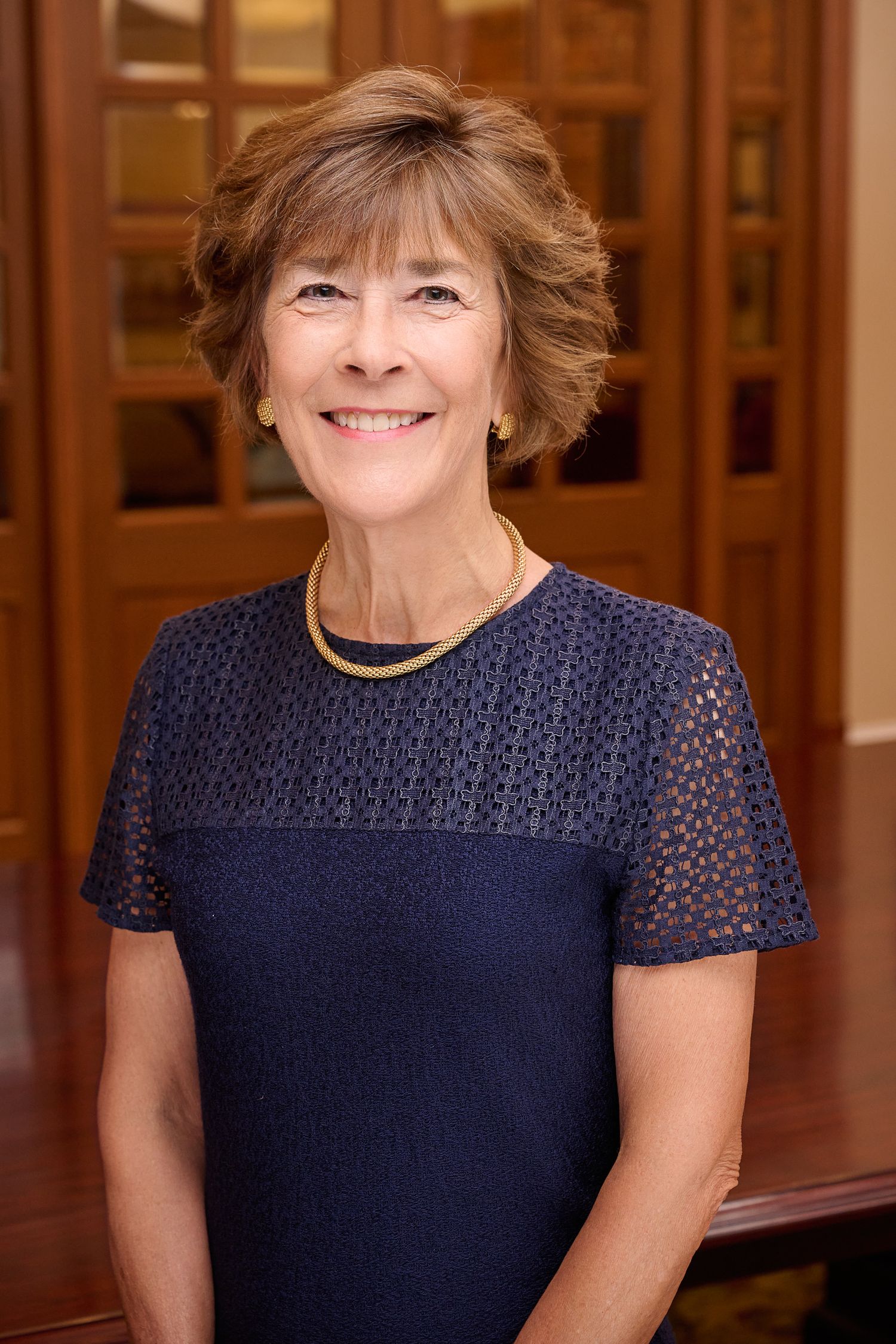 Professional woman in navy blue dress with gold necklace smiles warmly in elegant wood-paneled interior setting.