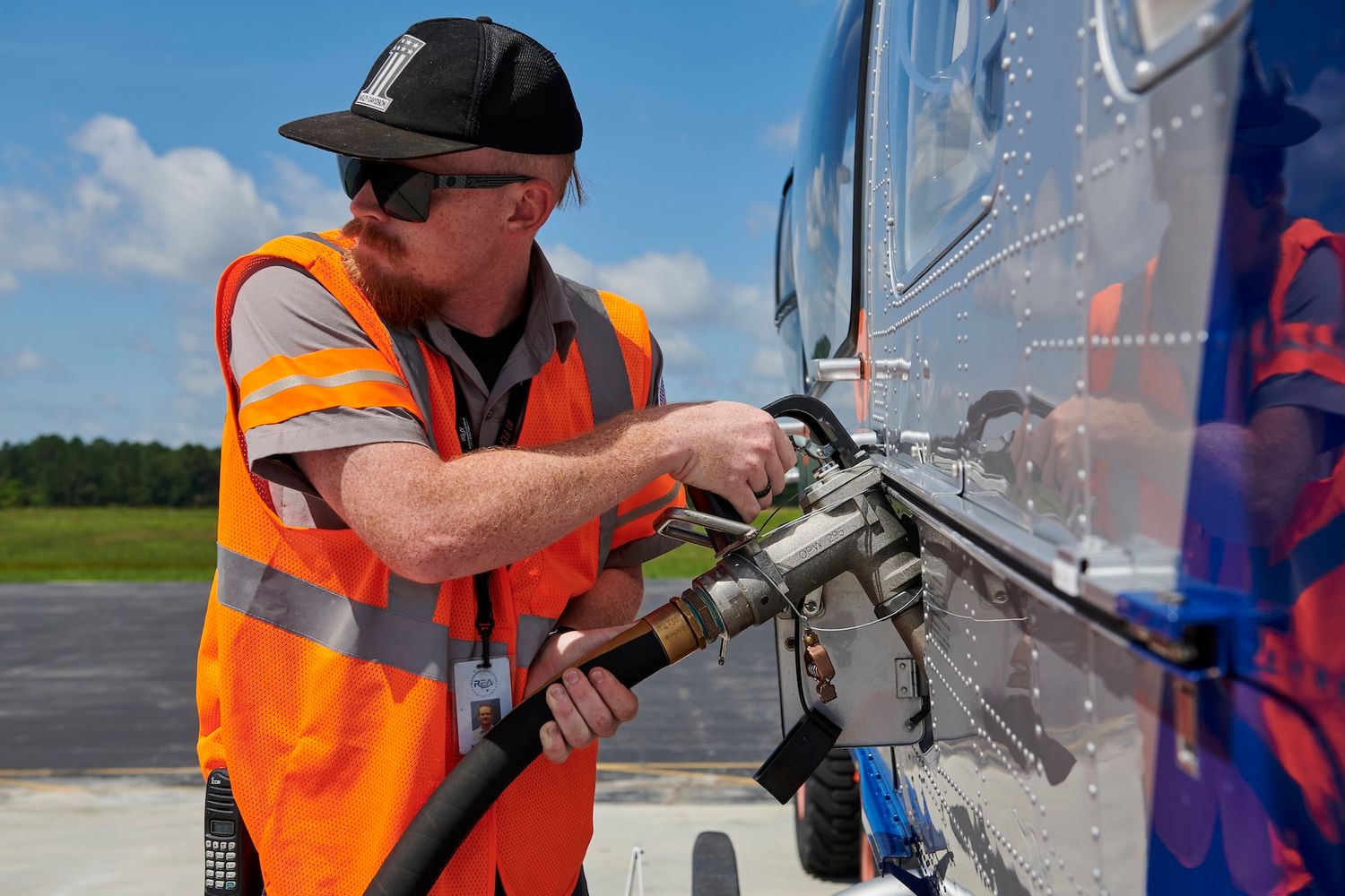 A worker in an orange safety vest refuels a helicopter on a sunny airport tarmac.