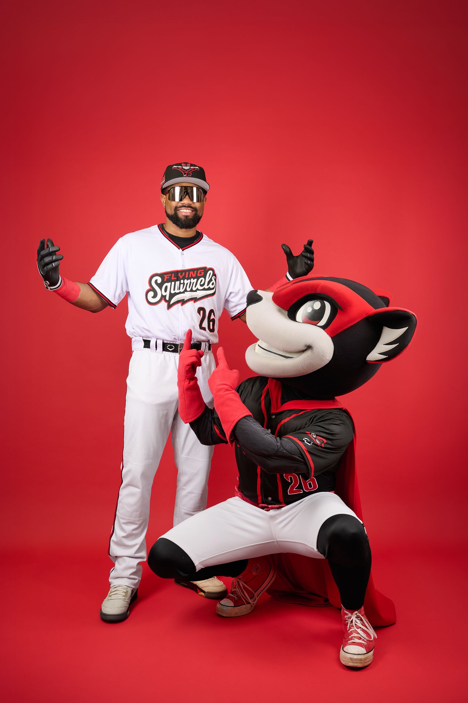 Baseball player in Richmond Squirrels uniform poses with team mascot against red background.