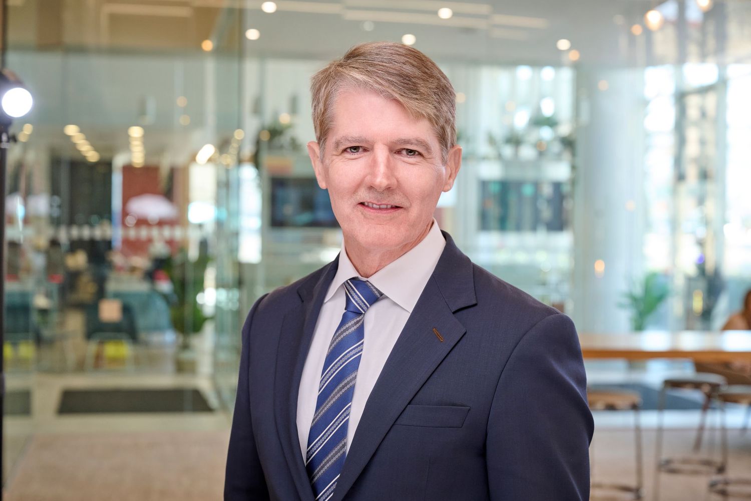 Senior businessman in navy suit and tie smiles in bright modern glass office building interior.