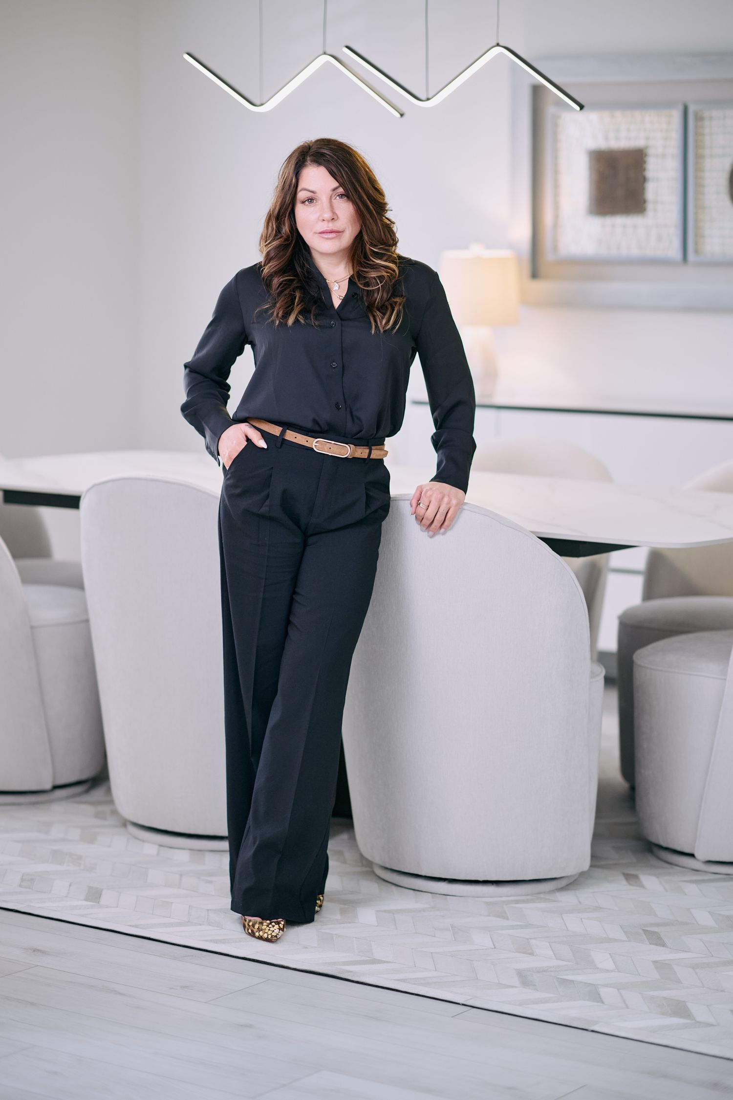 Professional woman in black outfit leaning against white chair in modern interior.