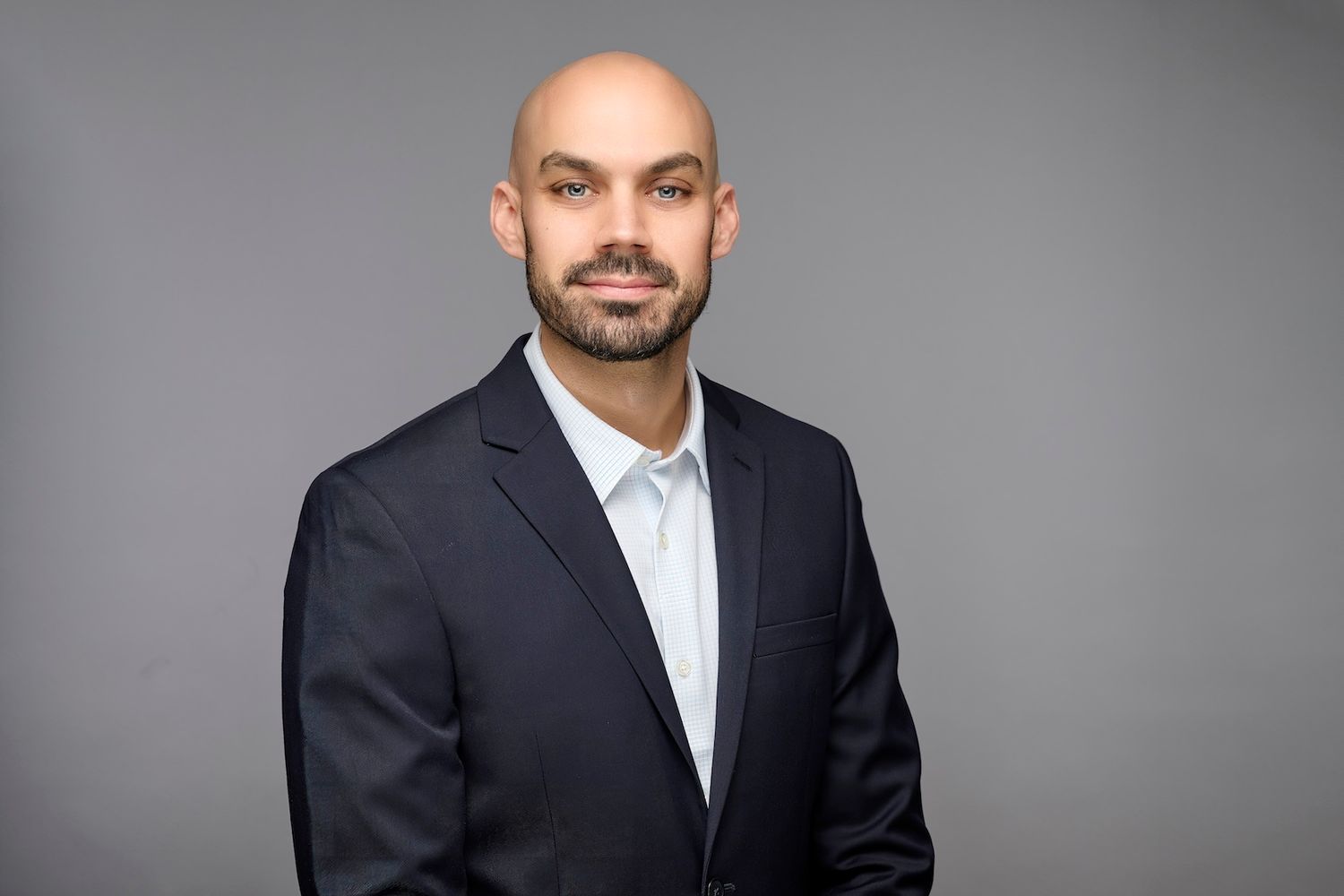 Professional headshot of bald man with beard wearing dark navy blazer on gray background.