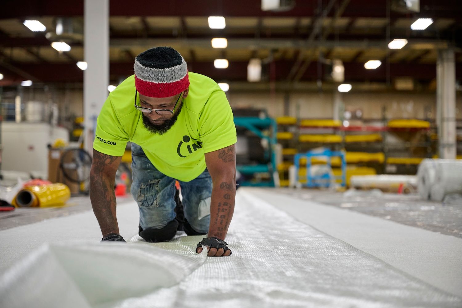 A worker in a neon shirt kneels on large white material in an industrial warehouse.