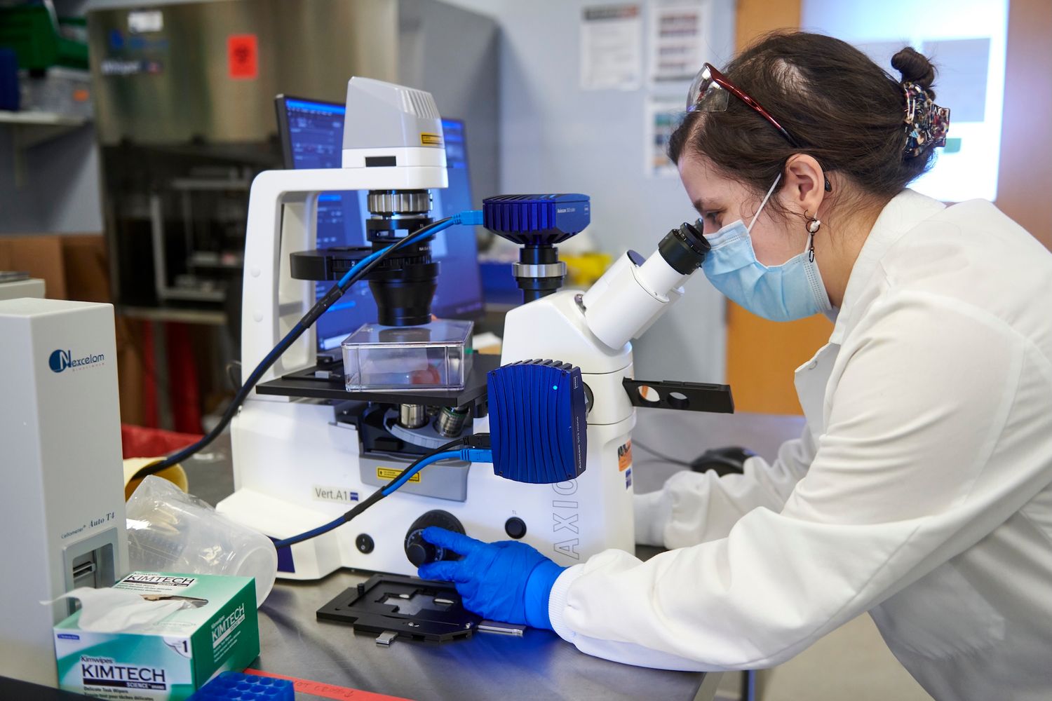 Female scientist in white lab coat and mask examining samples under a microscope in a laboratory.