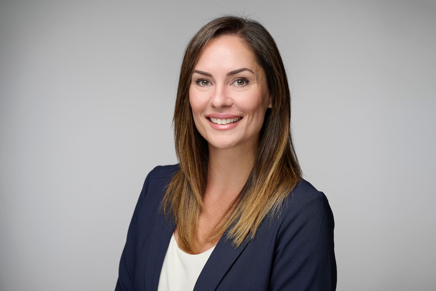 Professional woman with brown hair in navy blazer smiling against neutral gray background.