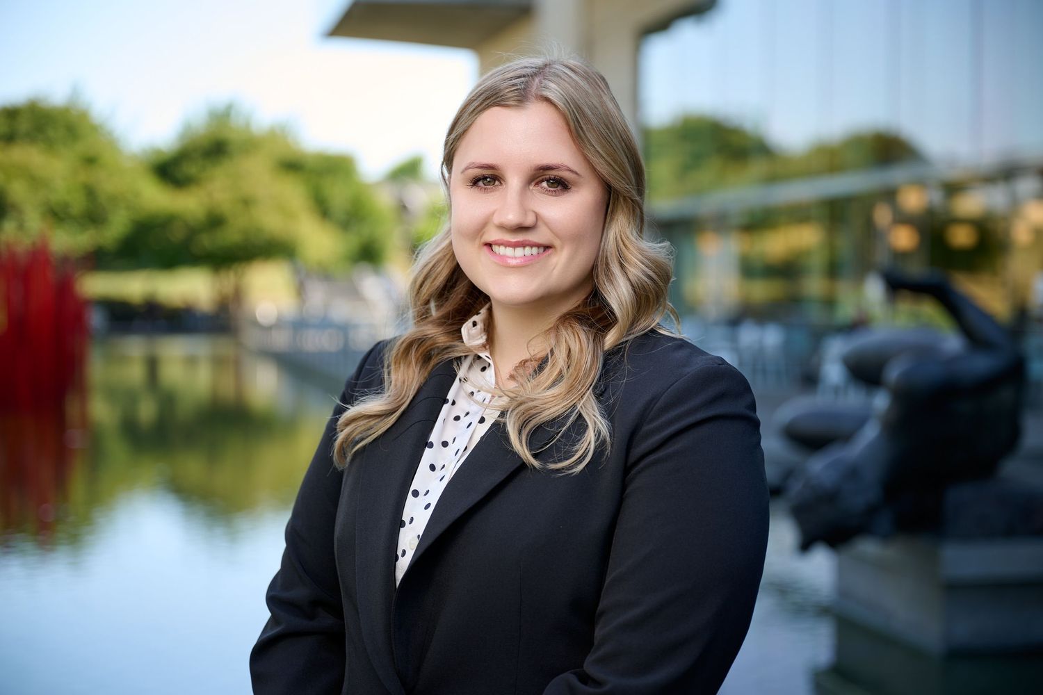 Young blonde professional woman in black blazer smiles outdoors near reflective water feature and sculpture garden.