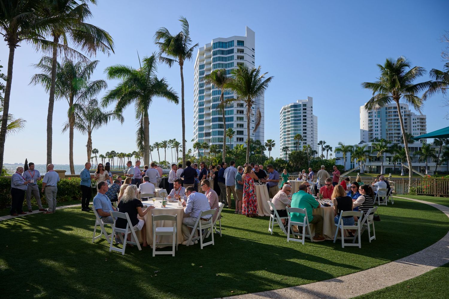 Outdoor waterfront event with guests dining on lawn surrounded by palm trees and luxury condos.