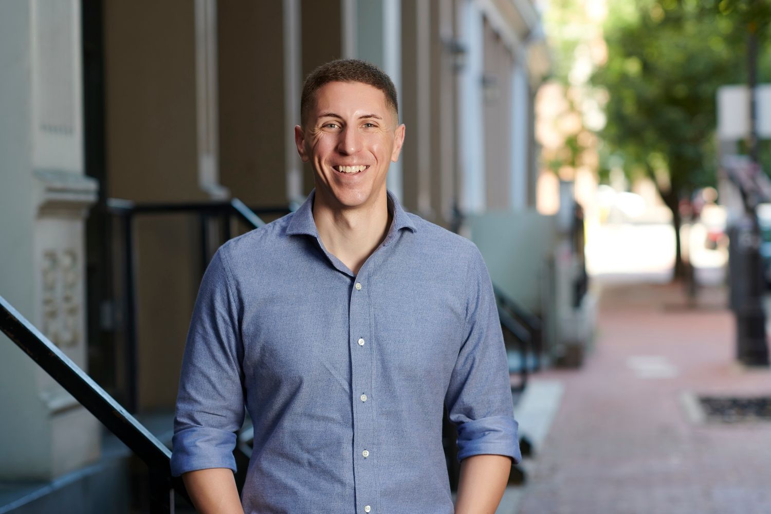 Smiling young man in blue shirt poses outdoors on urban brick sidewalk with buildings behind.