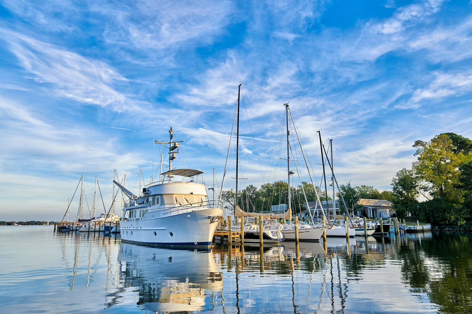 Sailboats and a white motor yacht docked at a calm marina reflecting in still water under a blue sky.