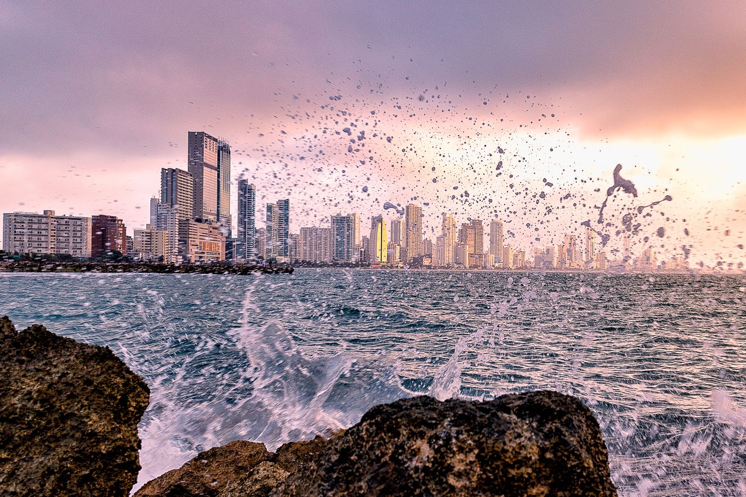 Ocean waves crashing on rocks with a city skyline and birds at sunset.