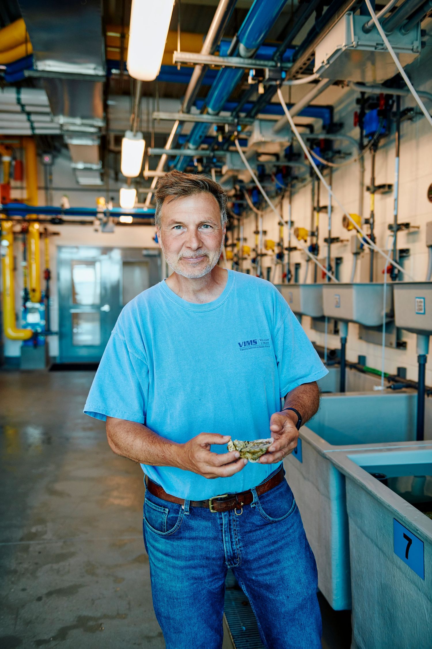 Middle-aged man in blue t-shirt holding a crab inside an aquaculture facility with tanks.