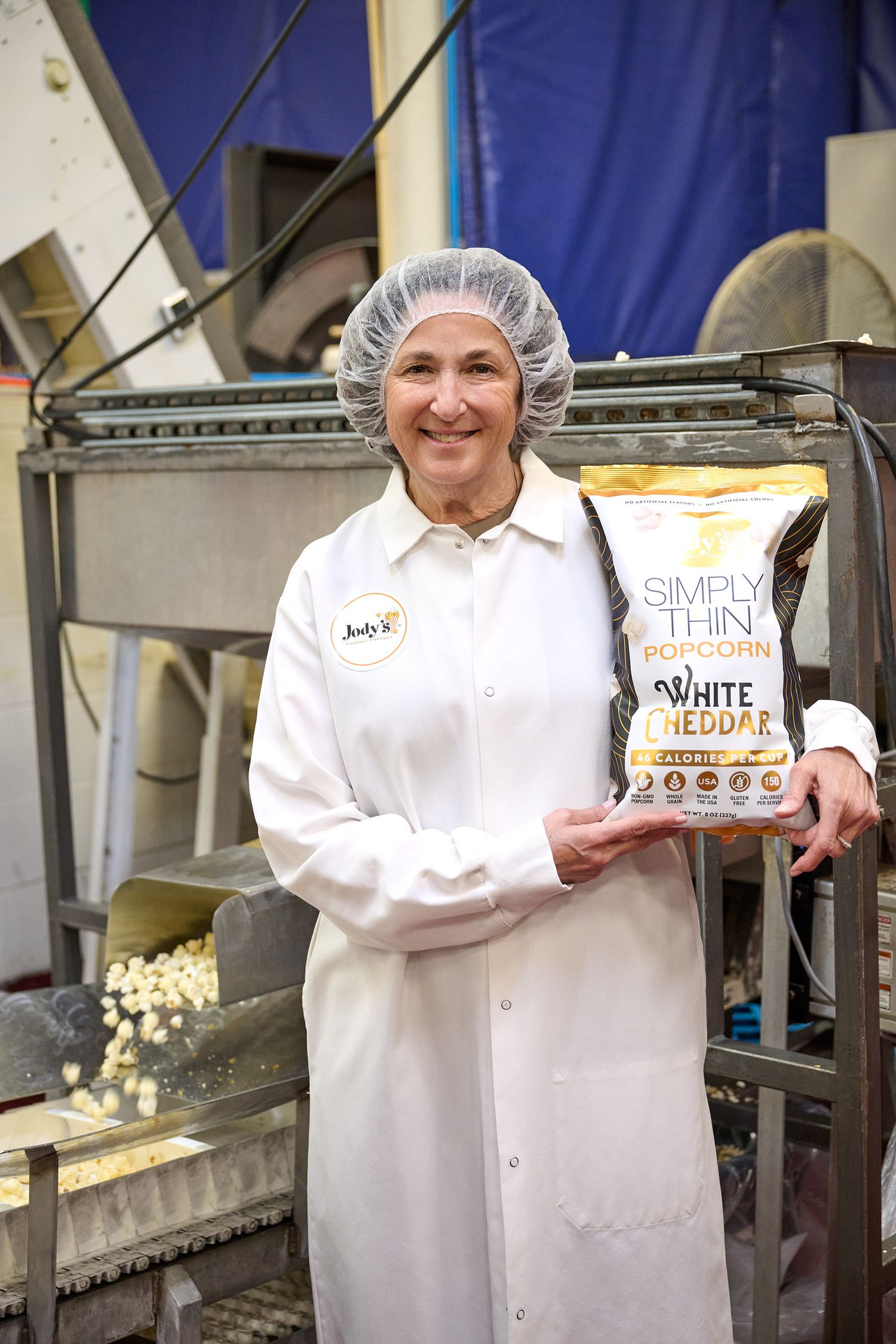 Smiling woman in a white lab coat and hairnet holding a bag of Simply Thin White Cheddar Popcorn.