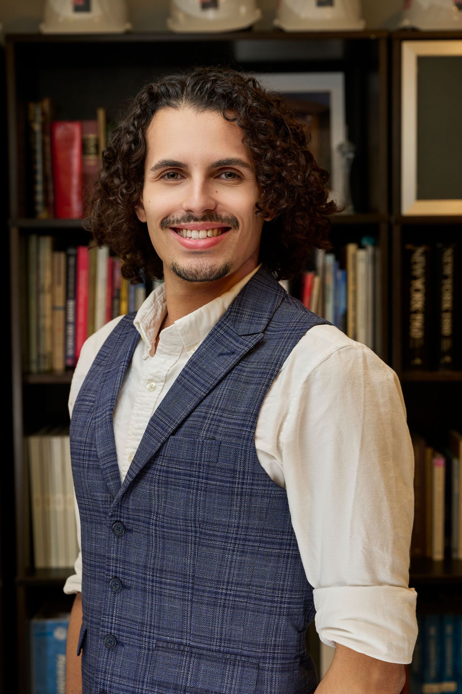 Young man with curly hair and a beard smiling in front of a bookshelf wearing a plaid vest.