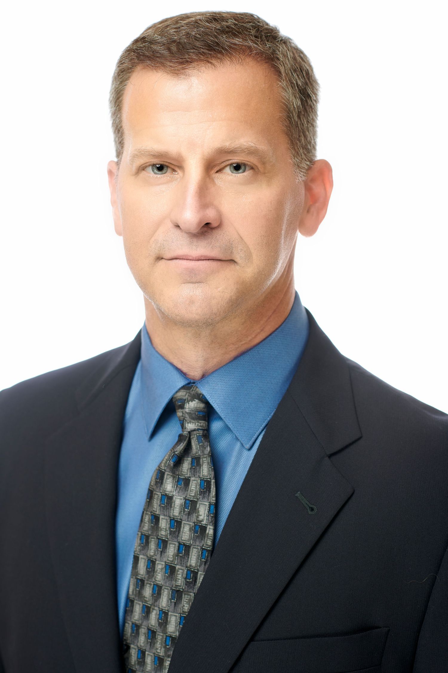 Professional headshot of a man in a dark suit, blue shirt, and patterned tie against a white background.