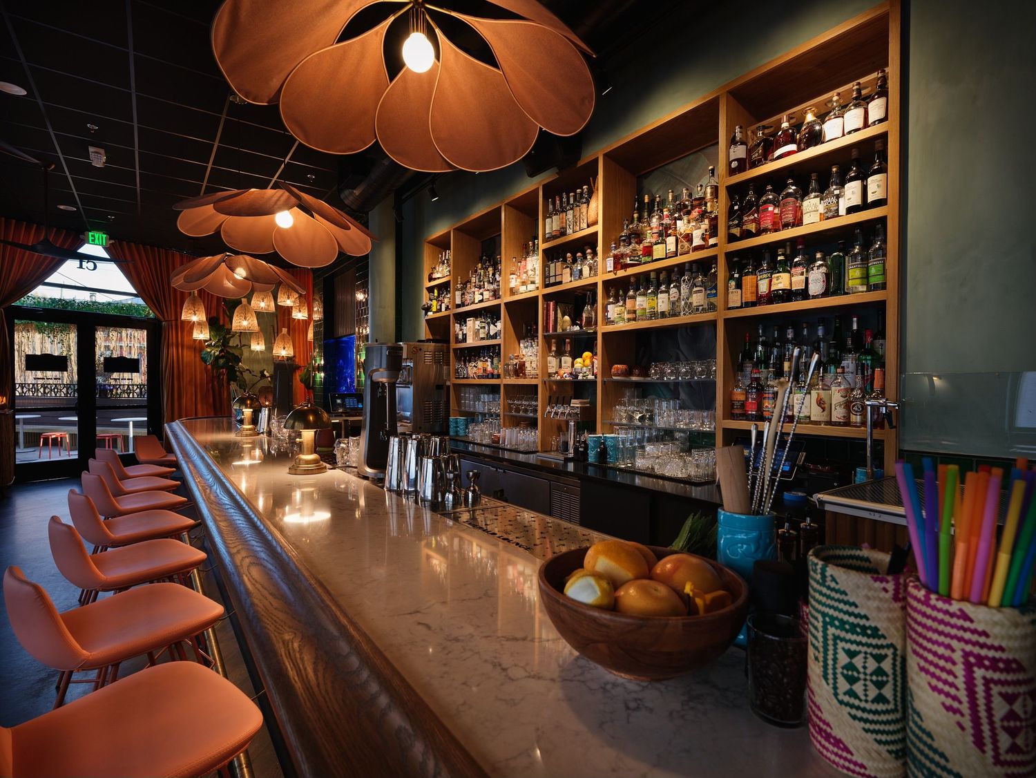 Vibrant tropical bar with long marble counter, orange pendant lights, and colorful bottle display.