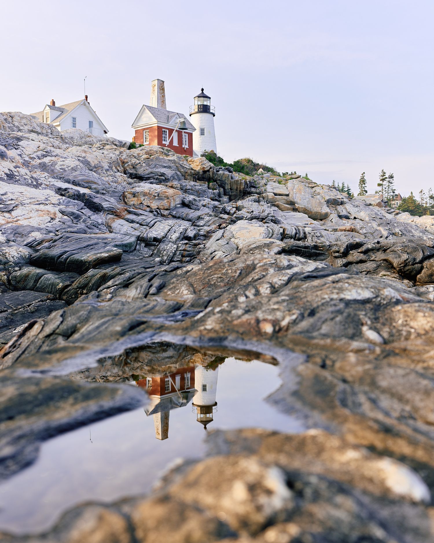 Historic lighthouse on rocky Maine coastline reflecting in tidal pool with red and white buildings nearby.