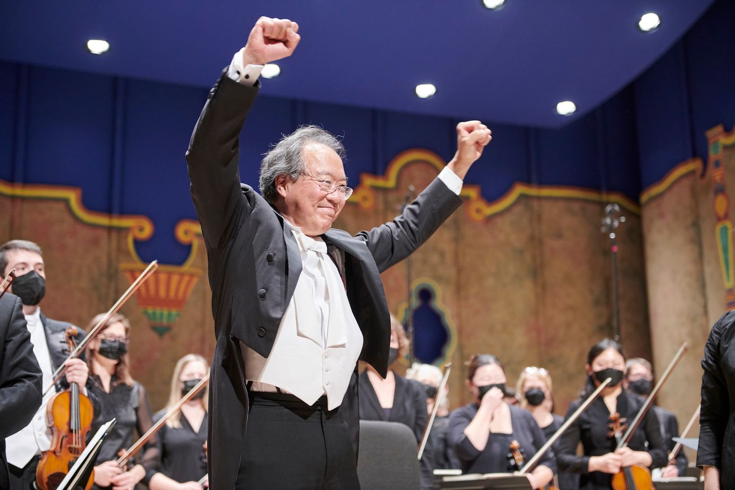 Orchestra conductor raising fists in triumph on stage before masked musicians.