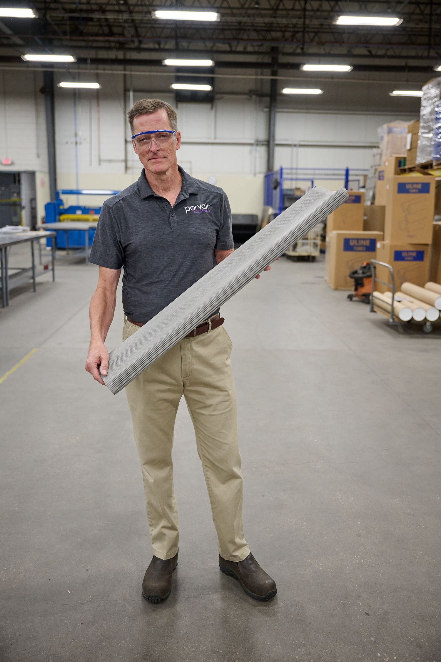 Man in safety glasses holds metal panel in large industrial warehouse facility.