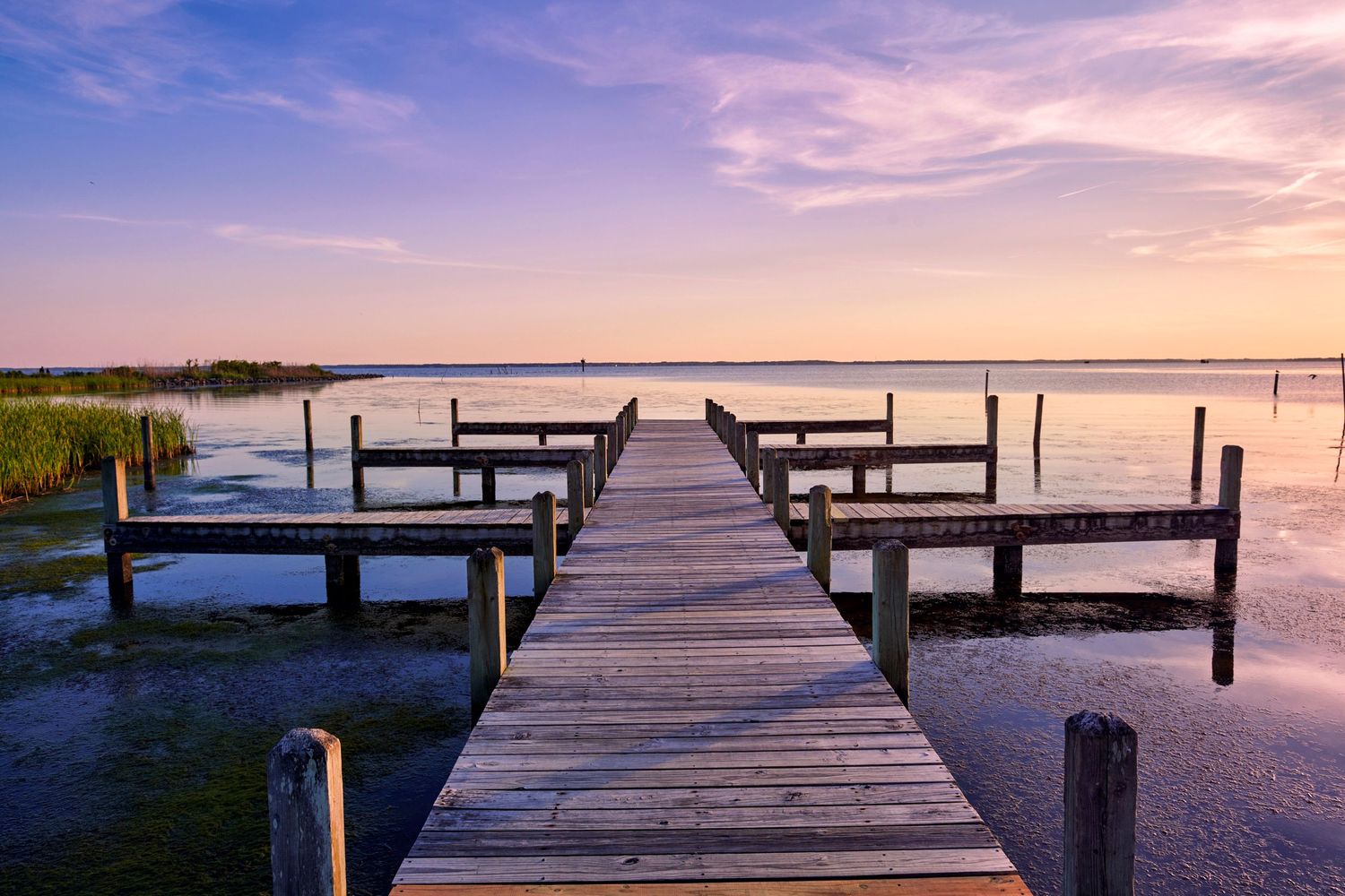 Wooden dock extending over calm waters at a serene pink and purple sunset.