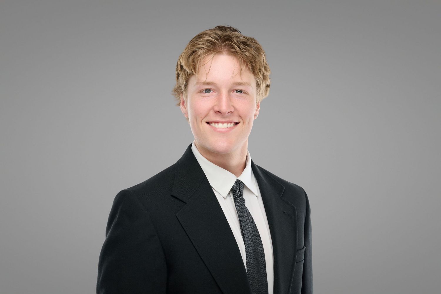 Young professional man in black suit and tie smiles against neutral gray background.