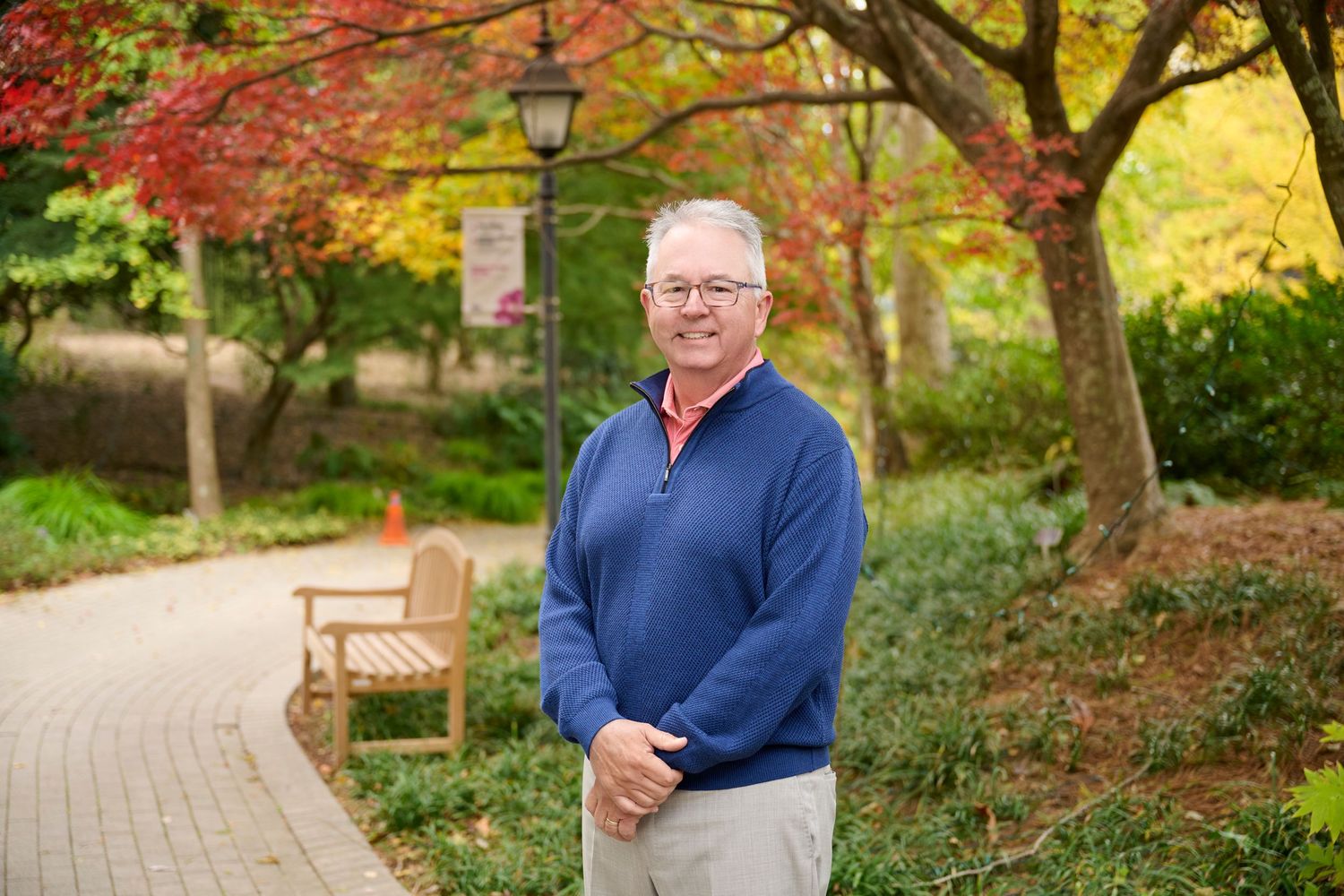 Smiling middle-aged man in blue pullover sweater stands on scenic autumn campus pathway with colorful foliage.