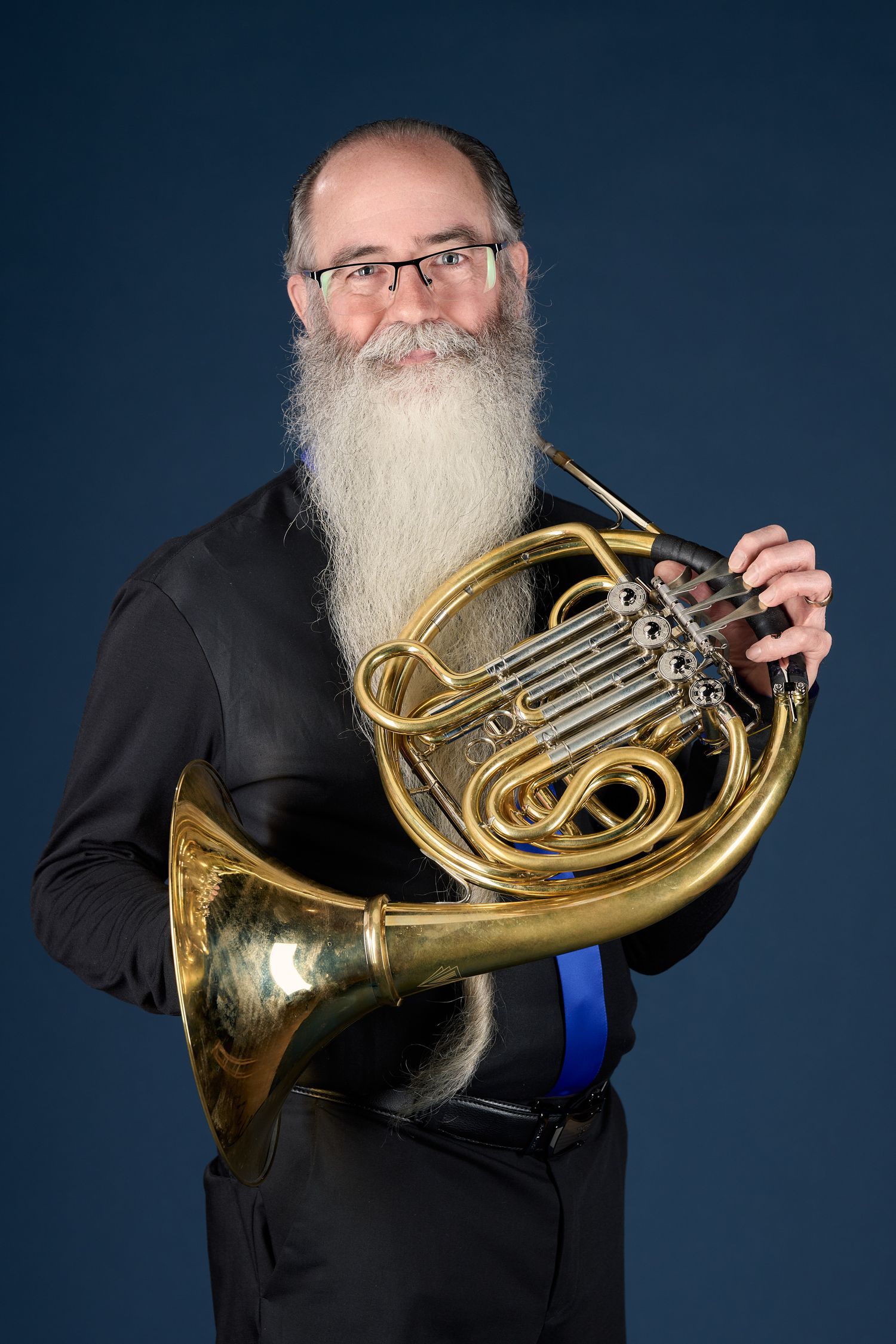 Bearded man with glasses holding French horn against blue background.