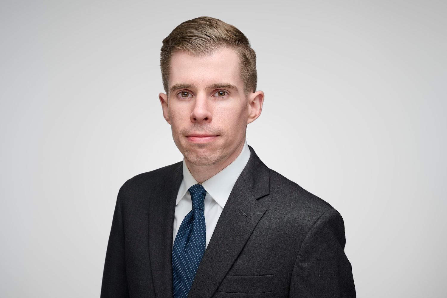 Young professional man in dark suit and blue tie poses for formal headshot on gray background.