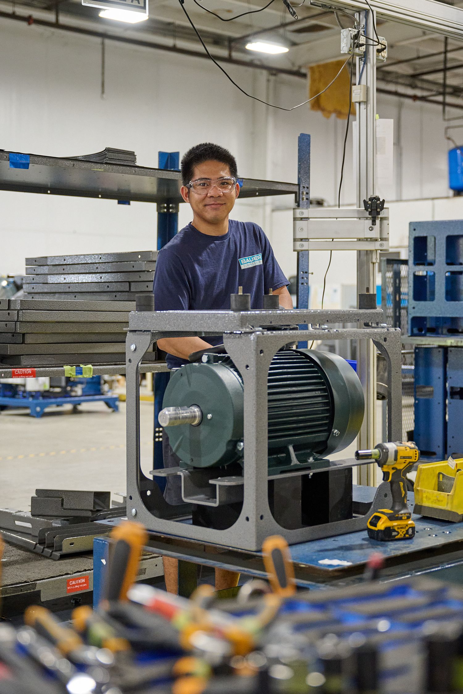 A smiling technician in safety glasses poses with an electric motor in a factory.