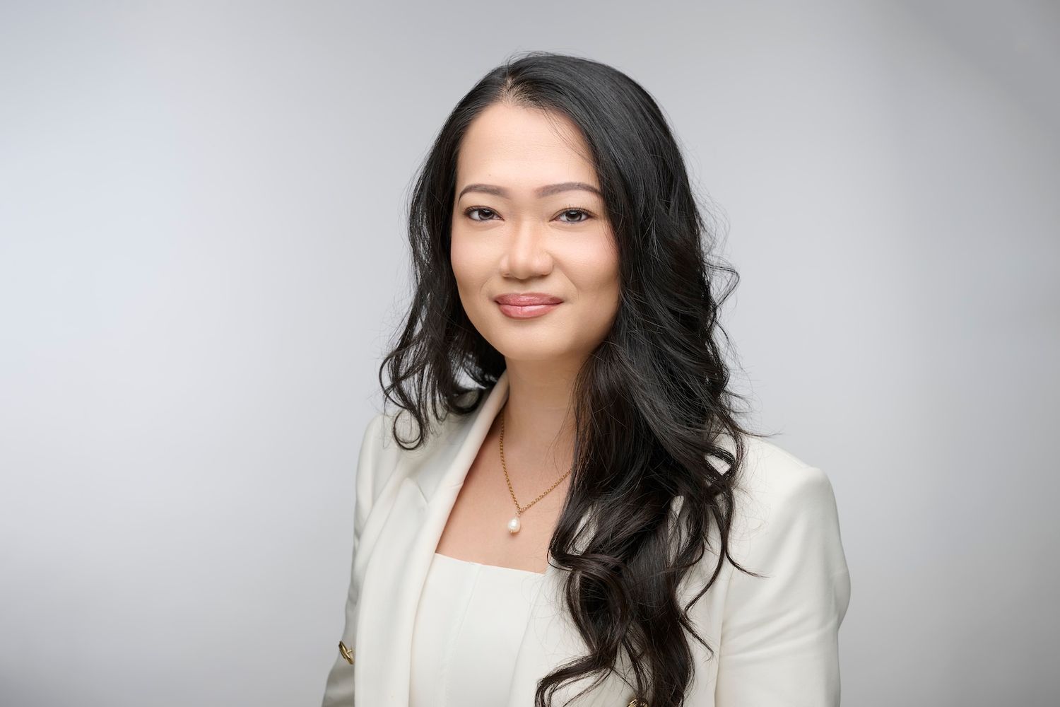 Professional Asian woman in white blazer with long dark hair poses against soft gray studio background.