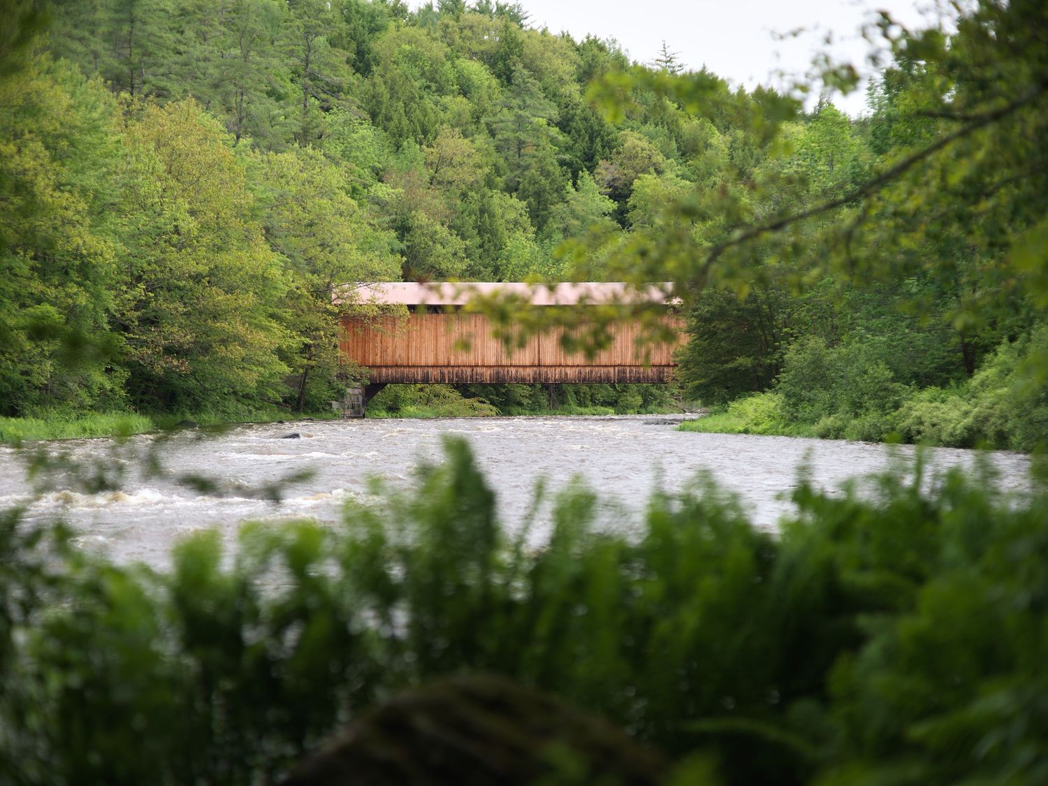 Historic red covered wooden bridge spanning a rushing river surrounded by lush green forest trees.