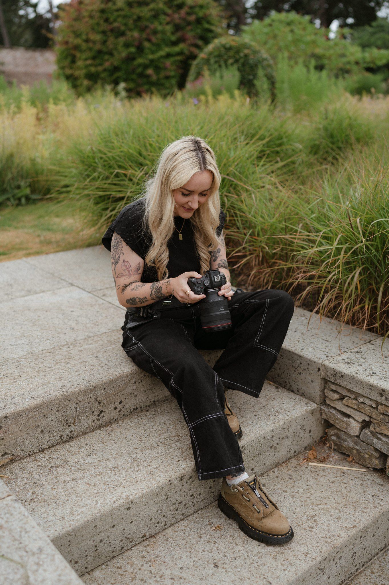 Blonde woman with tattoos sits on stone steps reviewing photos on her camera, surrounded by lush garden greenery.