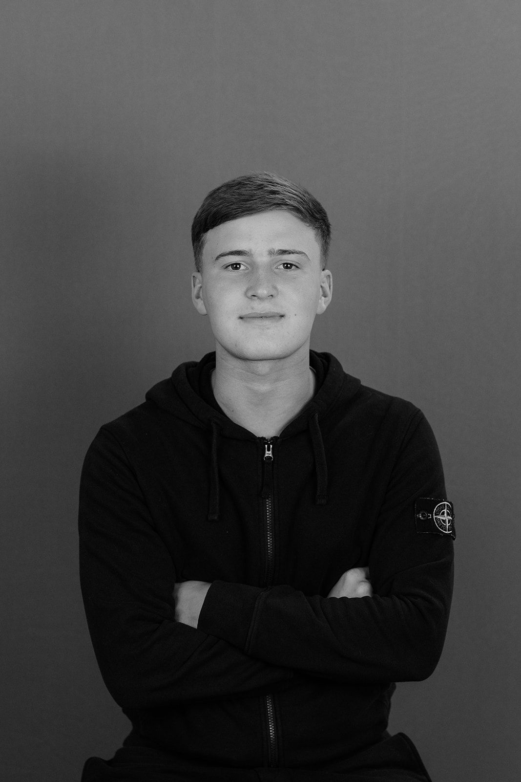 Black and white portrait of a young man in a Stone Island hoodie with arms crossed.