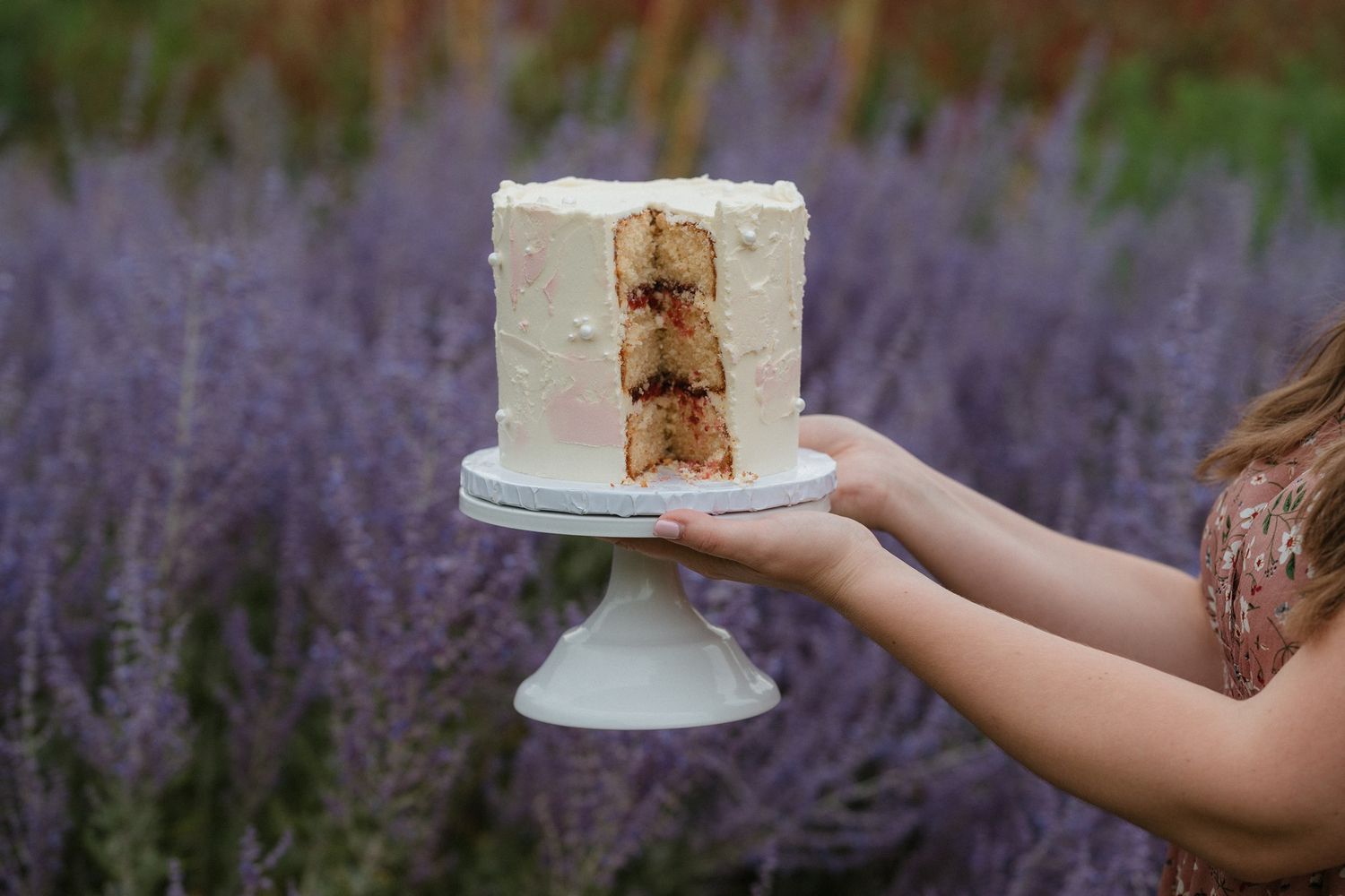 A sliced white frosted layer cake on a pedestal stand held outdoors against a purple lavender field backdrop.