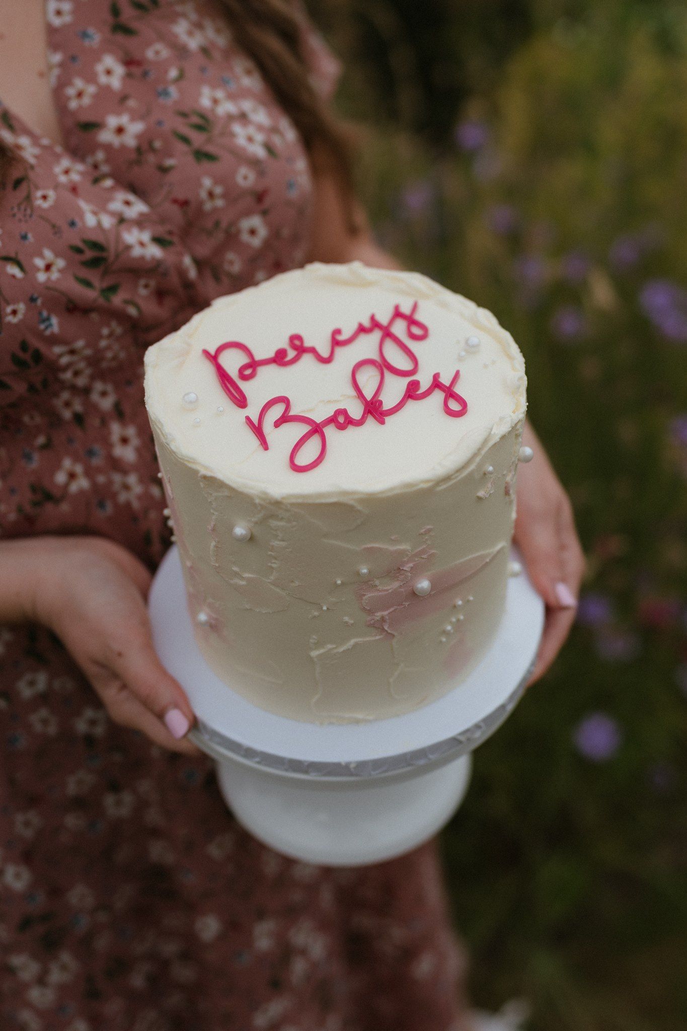 Woman holding a white cake stand with a cream frosted cake reading Percy's Baby in pink cursive frosting.