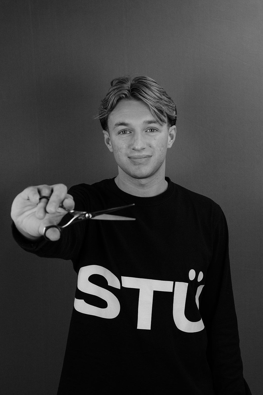 Young man in black STU sweatshirt holding scissors toward camera in black and white portrait.