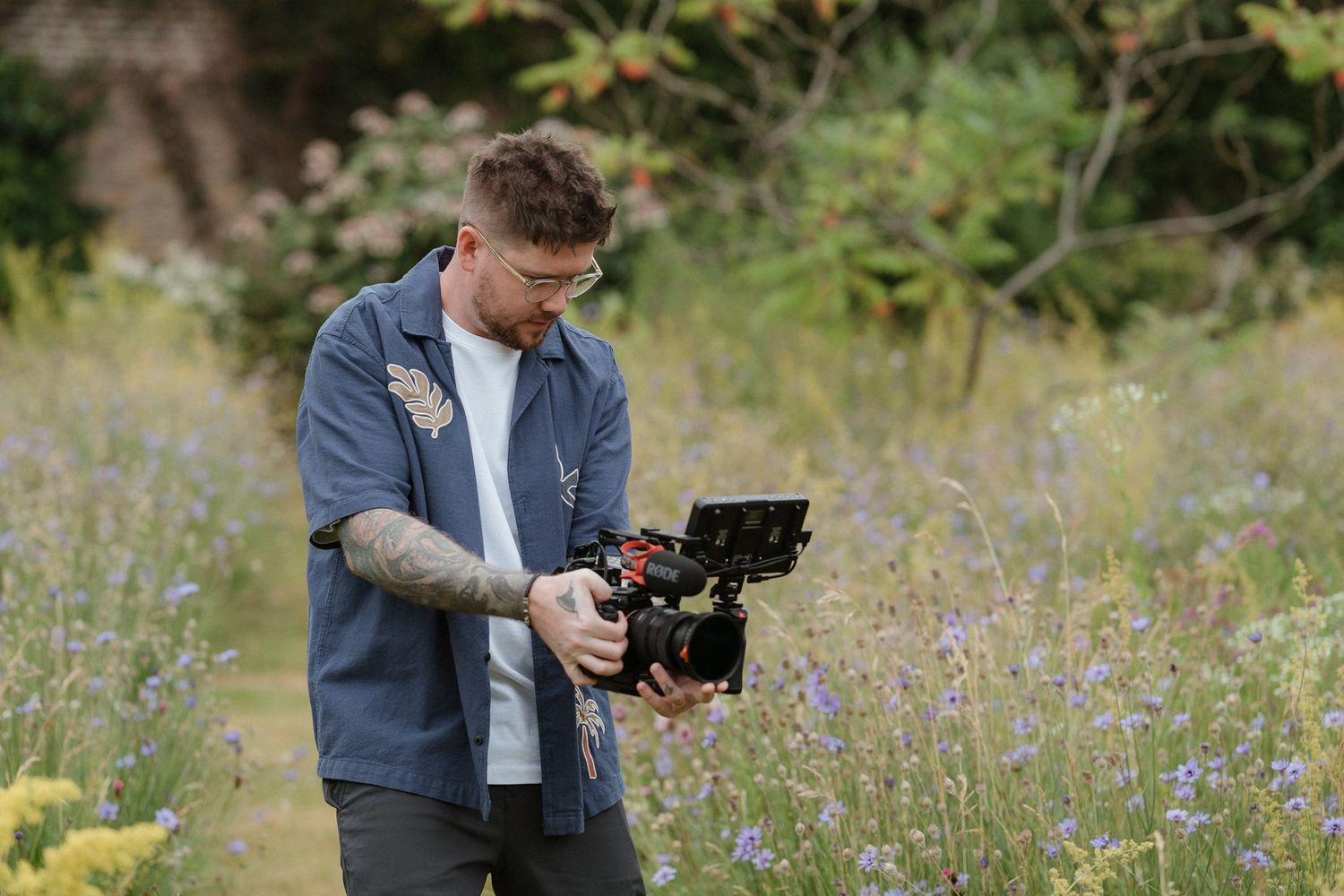 A tattooed male videographer holds a professional camera rig while filming outdoors in a wildflower meadow.