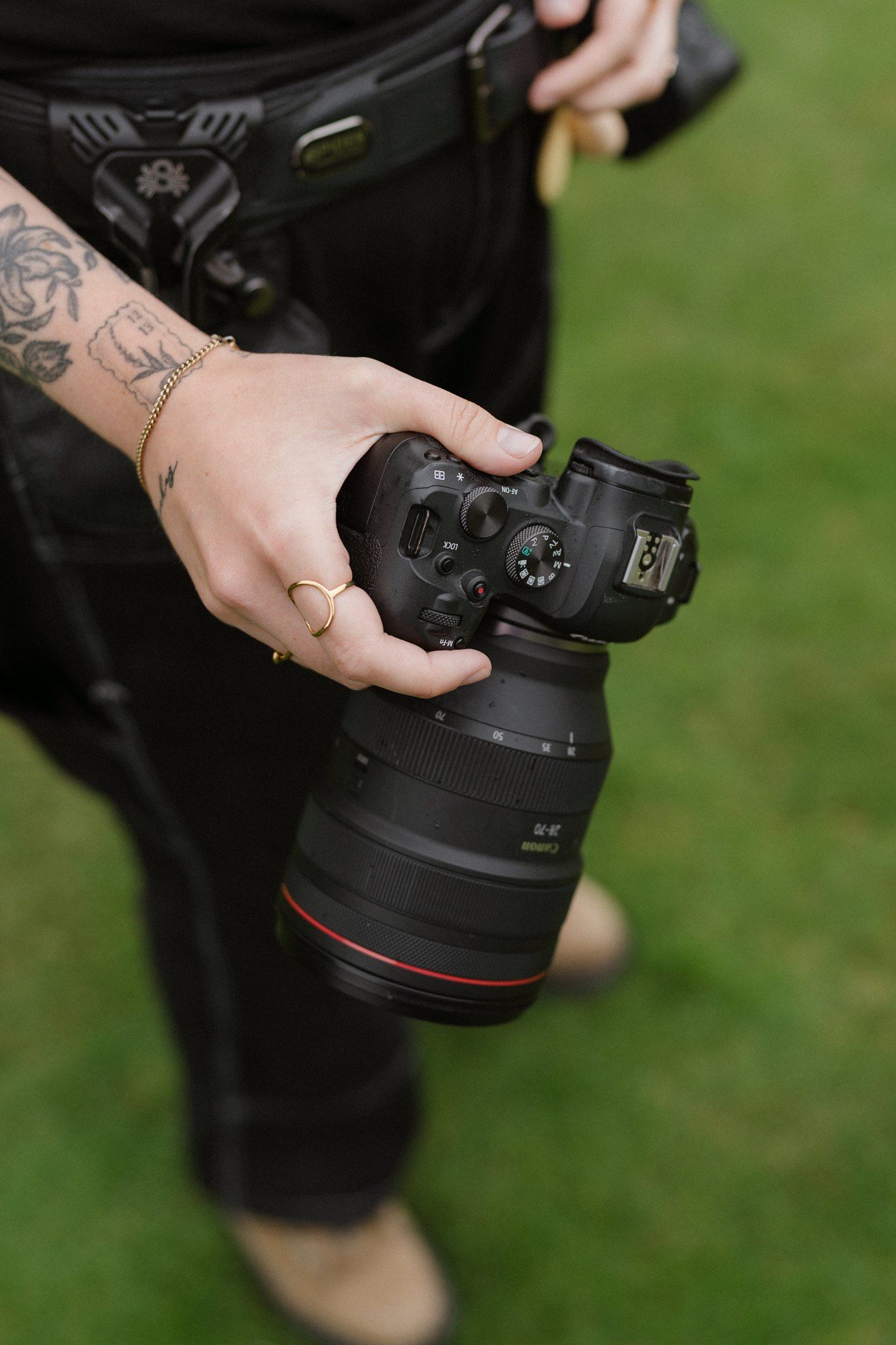 Tattooed hand holding a Canon DSLR camera with zoom lens outdoors on green grass background.