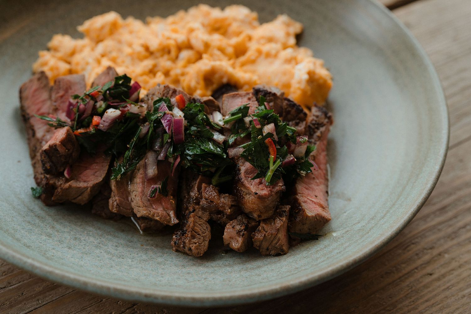 Sliced medium-rare steak topped with chimichurri sauce served with creamy mashed sweet potato on a plate.