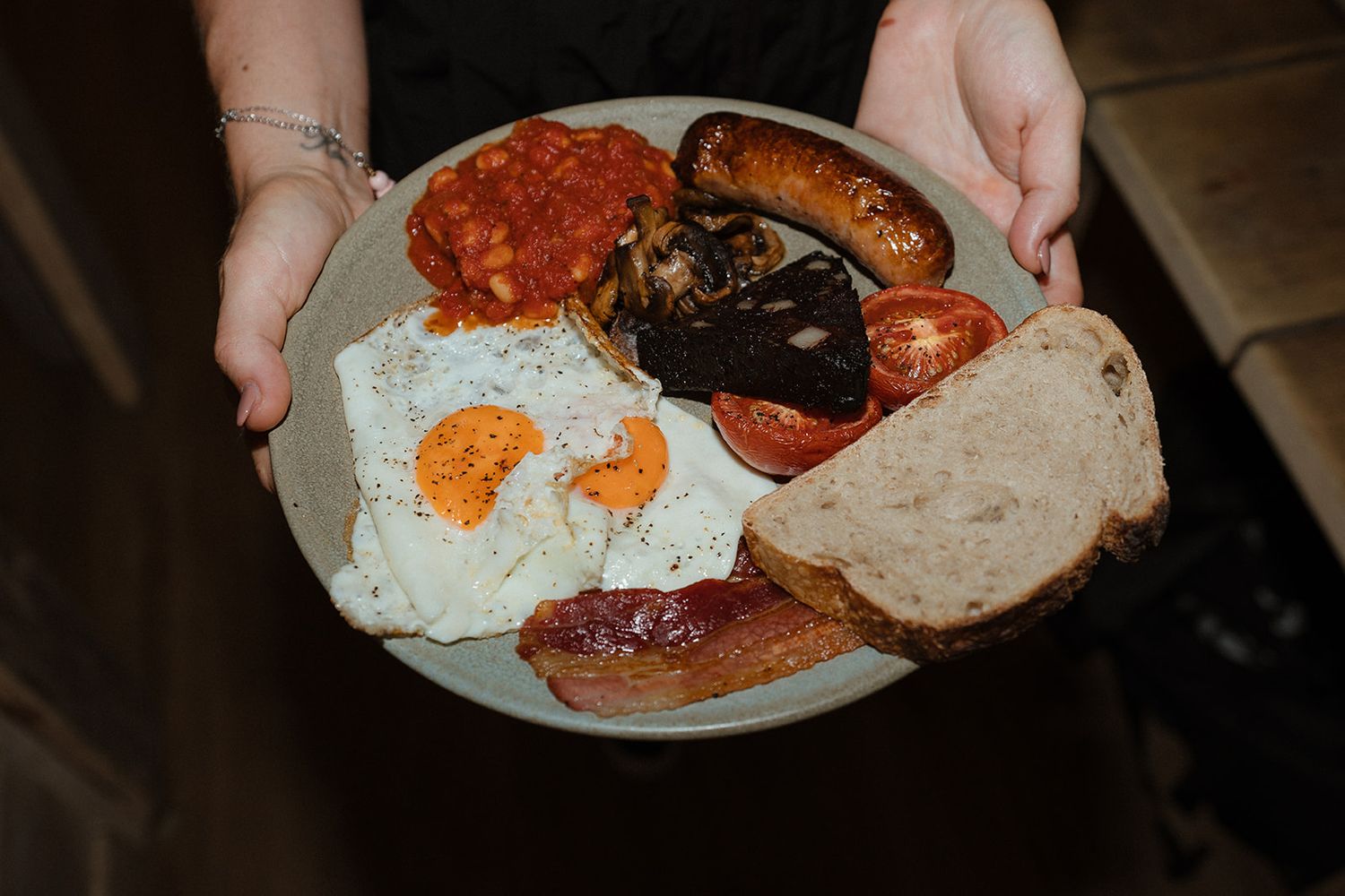 Hands holding a full English breakfast plate with fried eggs, bacon, sausage, tomatoes, and toast.